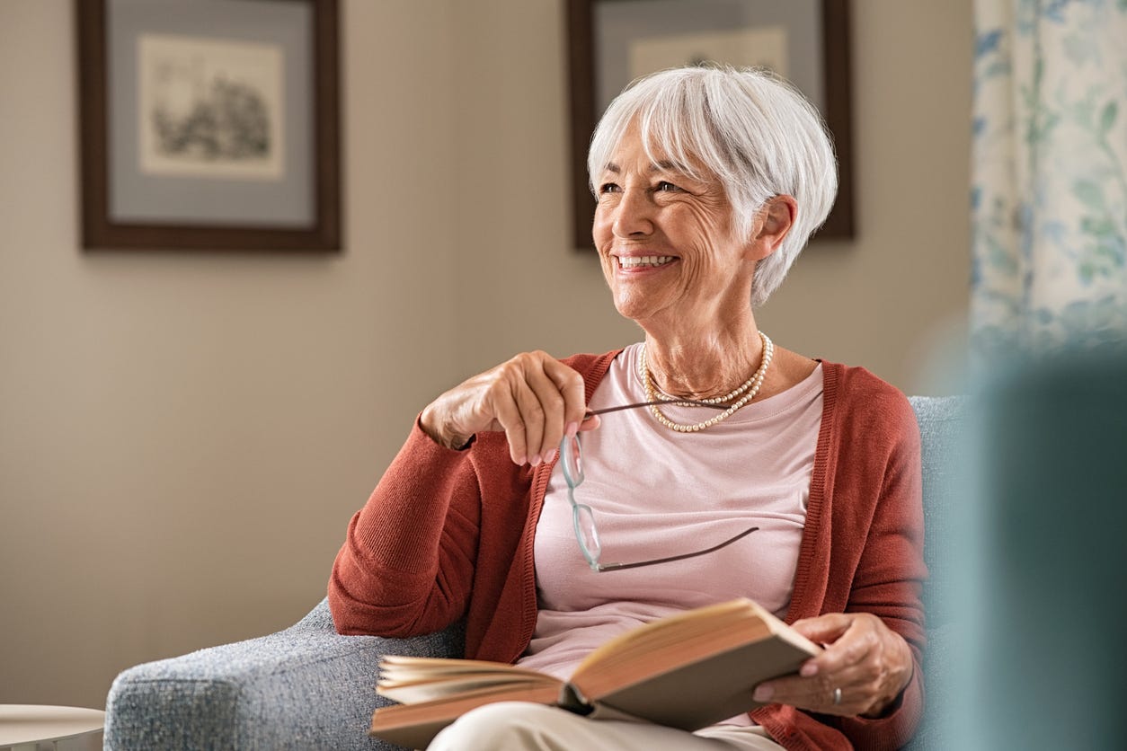 Cheerful senior woman holding book and eyeglasses thinking while relaxing at home. Happy elderly woman reading book at home sitting on couch. Beautiful old teacher takes a break from reading while looking through the window with a big grin.