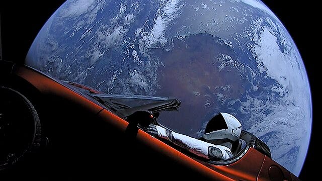 2018 yılında uzaya yollanmış Tesla arabanın fotoğrafı. Önde kırmızı araba ve içindeki bir astronot mankeni görünüyor. Arkada da kocaman, yuvarlak, mavi dünya.