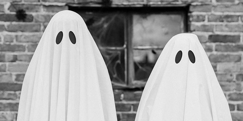 Two people in ghost sheet costumes stand in front of an old brick wall
