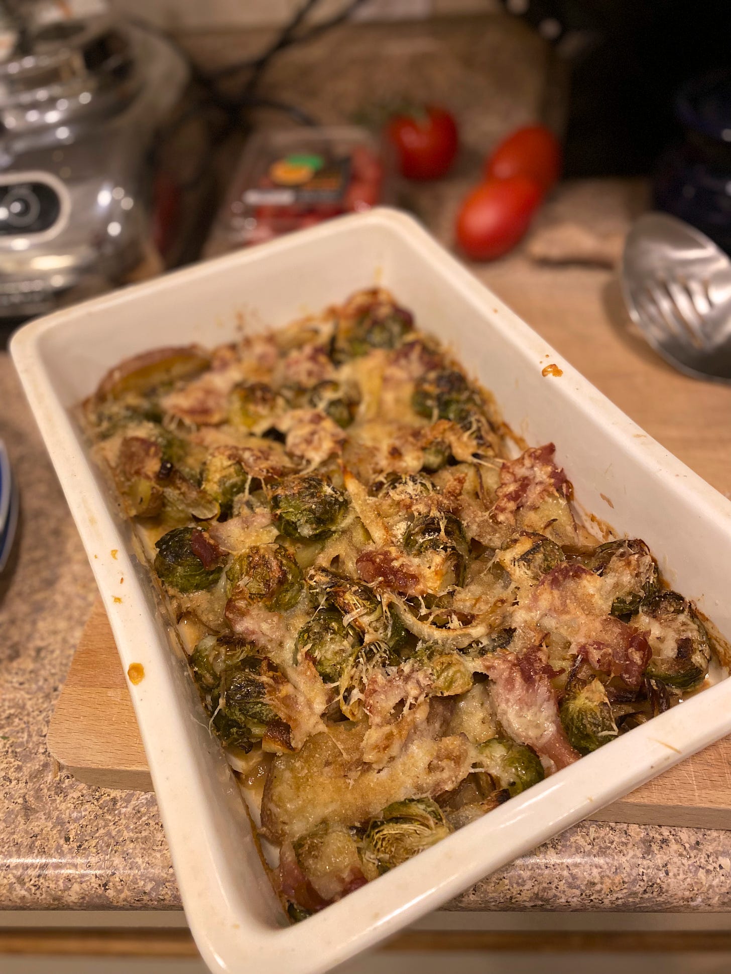 A white rectangular baking dish of the gratin described above. The cheese is browned in places and crisp pieces of prosciutto are visible throughout.