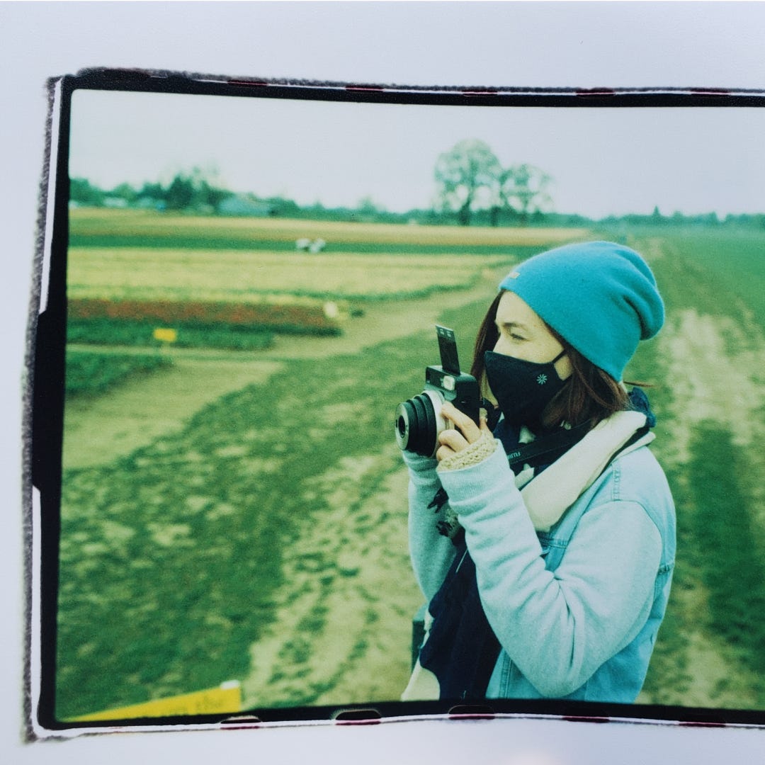 Sarah stands in a green field holding her camera. She is looking off into the distance, poised to take a photo. She wears a blue beanie, jean jacket, and a face mask.