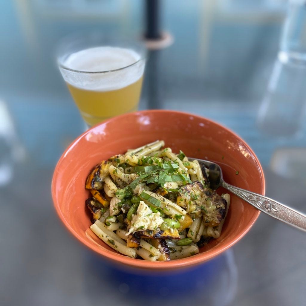 An orange bowl of casarecce in basil oil with charred pieces of yellow zucchini, torn chunks of fresh mozza, edamame, and capers. The top has ground black pepper and chopped basil. In the background is a glass of hazy pale ale.