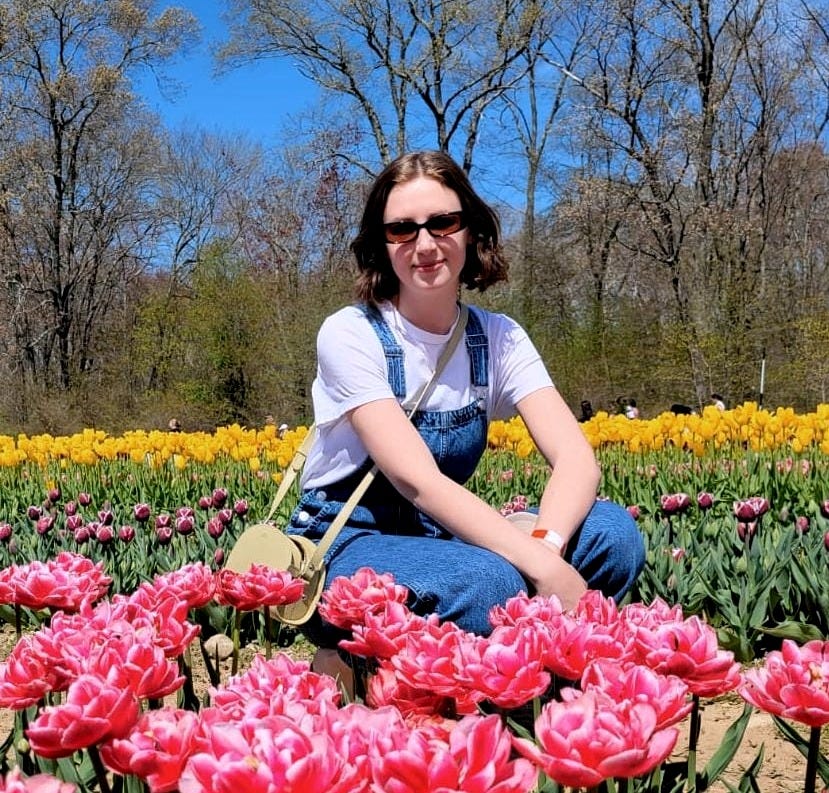 Aleksa, wearing jean overalls, a white t-shirt, black sunglasses and a green bag, kneels between rows of yellow, pink and purple tulips at a flower farm in Rhode Island.