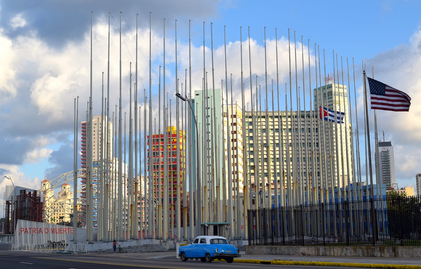 A blue car drives past a number of tall poles, a large American flag, and a smaller Cuban one. A number of colorful skyscrapers stand in the distance beneath a partially cloudy sky.