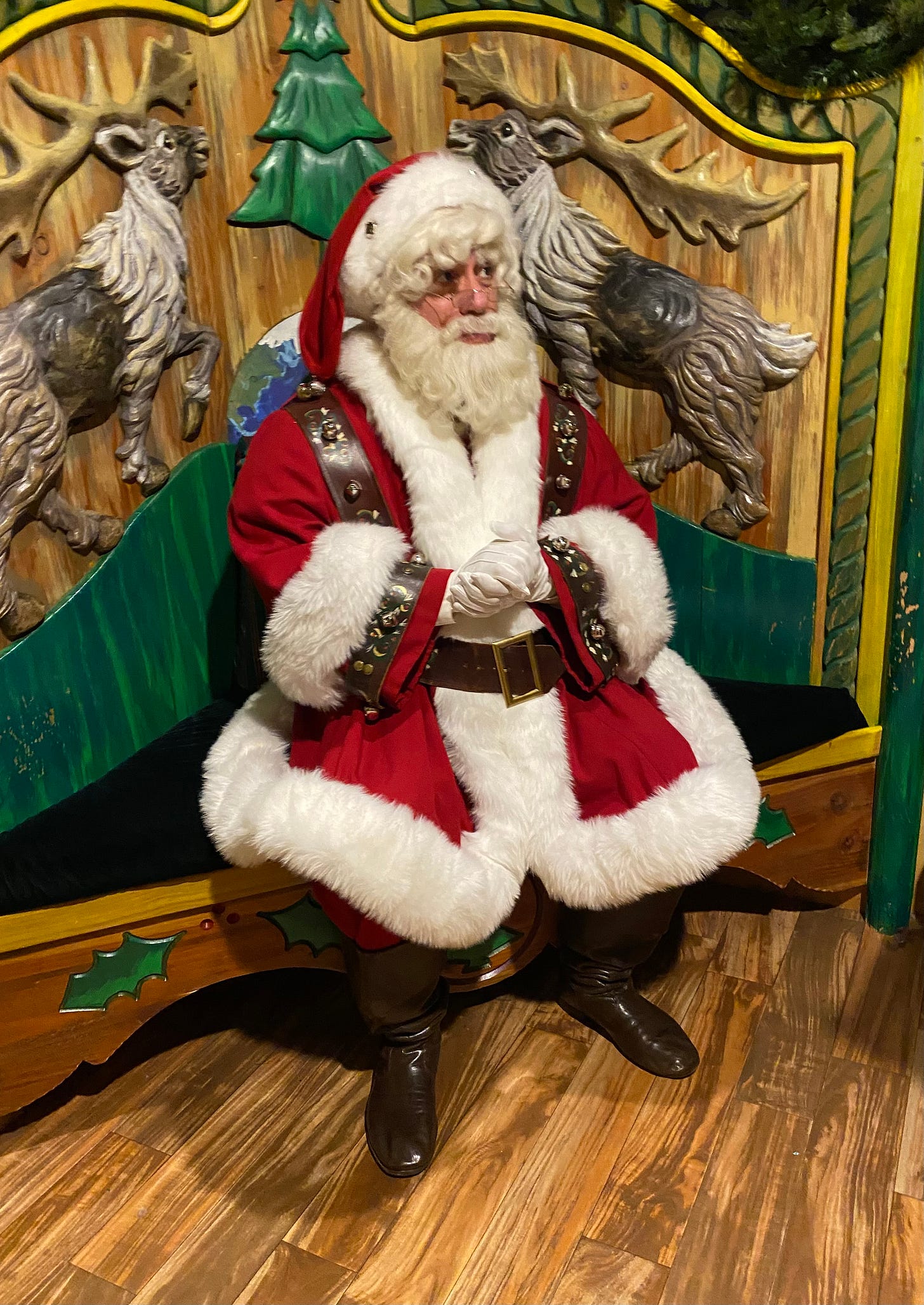 A man in a full Santa costume sits on a festively carved wooden bench.