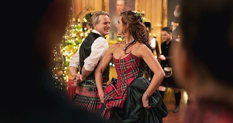 Movie still from A Castle for Christmas. A middle-aged couple dance in a lavish ballroom with Christmas lights, dressed in a kilt and a tartan dress.