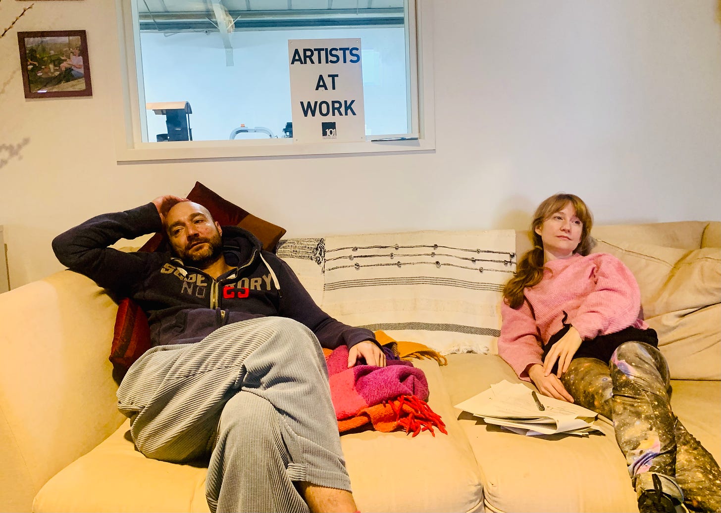 A man and a woman lie on a sofa. Behind them is a sign which reads "Artists at work"
