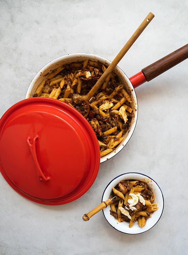 A large red pan with a dark wooden handle is filled with rigatoni pasta with red sauce and bits of mozzarella, there is a wooden spoon poking outside of the pan and there is a red lid half covering it. There is a small white bowl with blue rim filled with more pasta off to the bottom of the photo, with a bamboo handle sticking out of it. All are placed on a cement backdrop.
