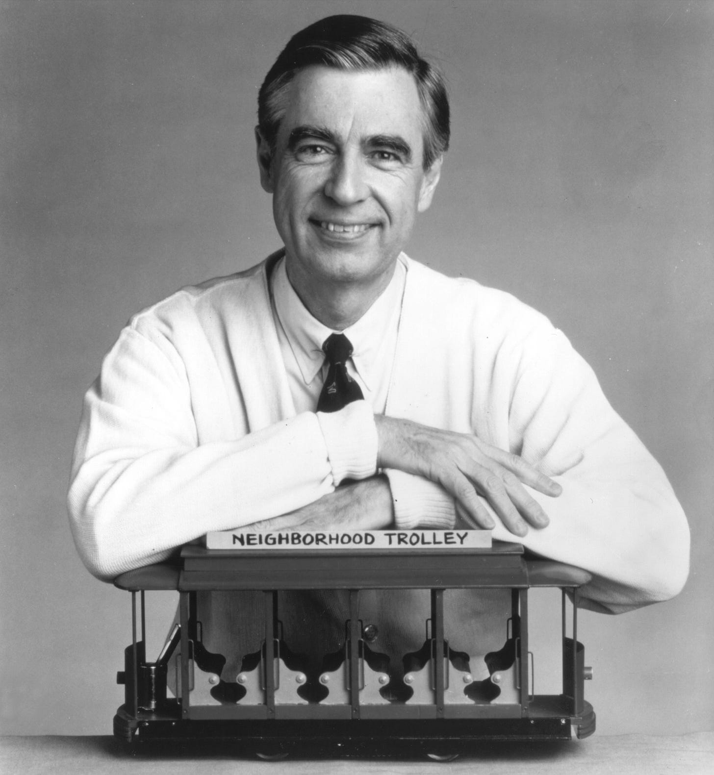 image of Mr. Rogers in a cardigan, leaning on the little trolley from his show