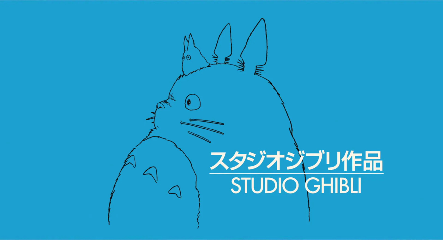 Logo do Studio Ghibli. Imagem com um fundo azul claro. Em branco, na parte direita inferior, escrito grande Studio Ghibli em inglês e em japonês, separados por uma linha. Desenhado ao fundo, em contorno preto, a imagem de Totoro e uma outra criatura da floresta: Totoro é um animal grande e peludo, como uma mistura de urso com gato. Ele tem orelhas pontudas, um corpo grande, redondo e peludo, grandes bigodes, um nariz pequeno e um contorno de barriga com três texturas como setas para cima. Em sua cabeça, o animal é pequeno, do tamanho da orelha. Ele é uma criatura com duas orelhas pontudas e dois olhos. Sem nenhum outro detalhe.