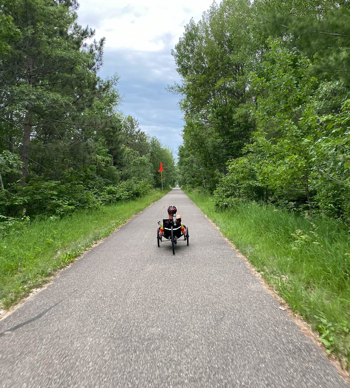 Biker on recumbant tricycle on straight paved trail with trees on either side.