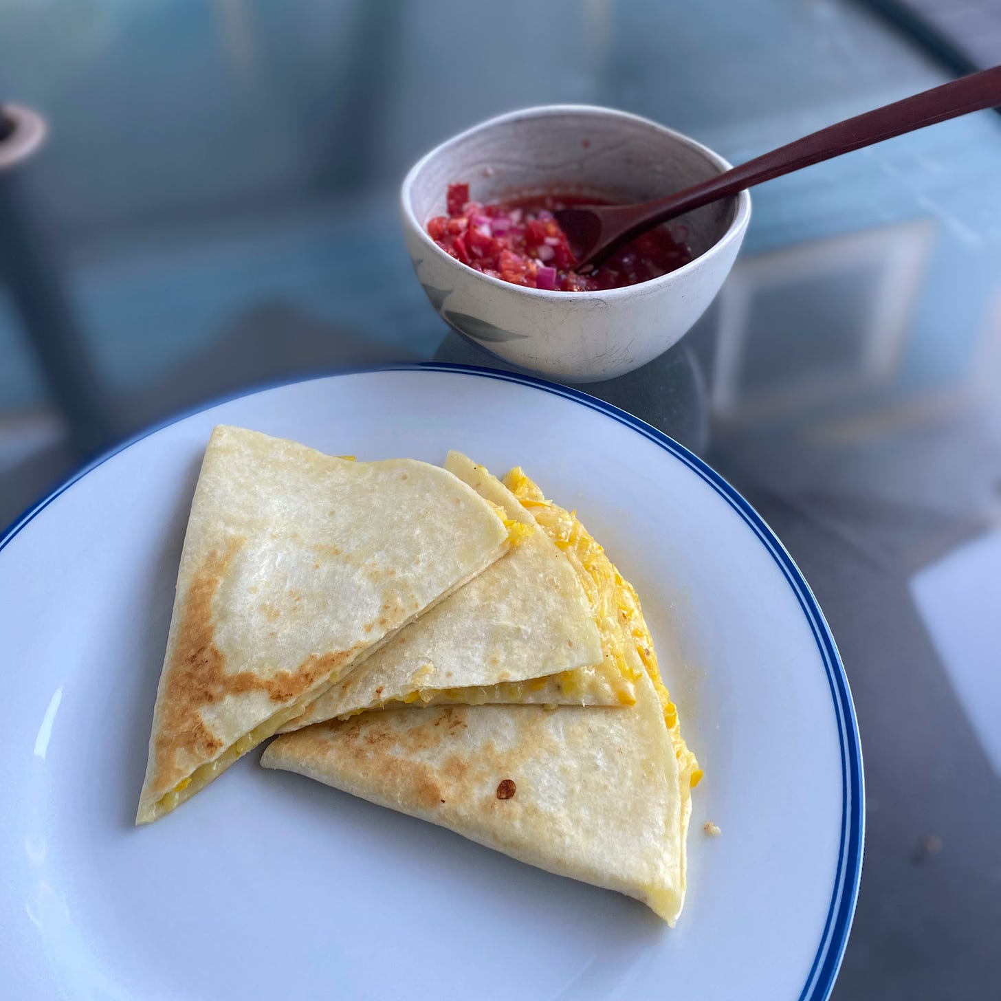 Three quesadilla triangles on a large white plate with a blue rim. Yellow zucchini and melted cheese are visible at the edge, and the tops are browned from the pan. In a small bowl behind the plate is the red onion and heirloom tomato salsa, with a spoon sticking out of it.