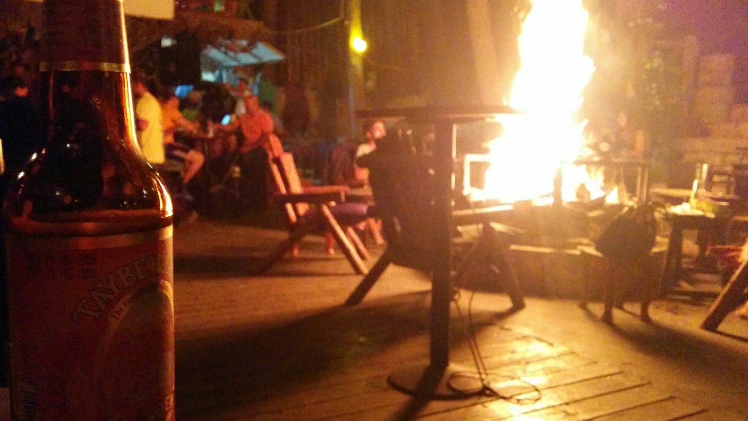 People sit in deck chairs drinking beer around a large fire at an outdoor bar