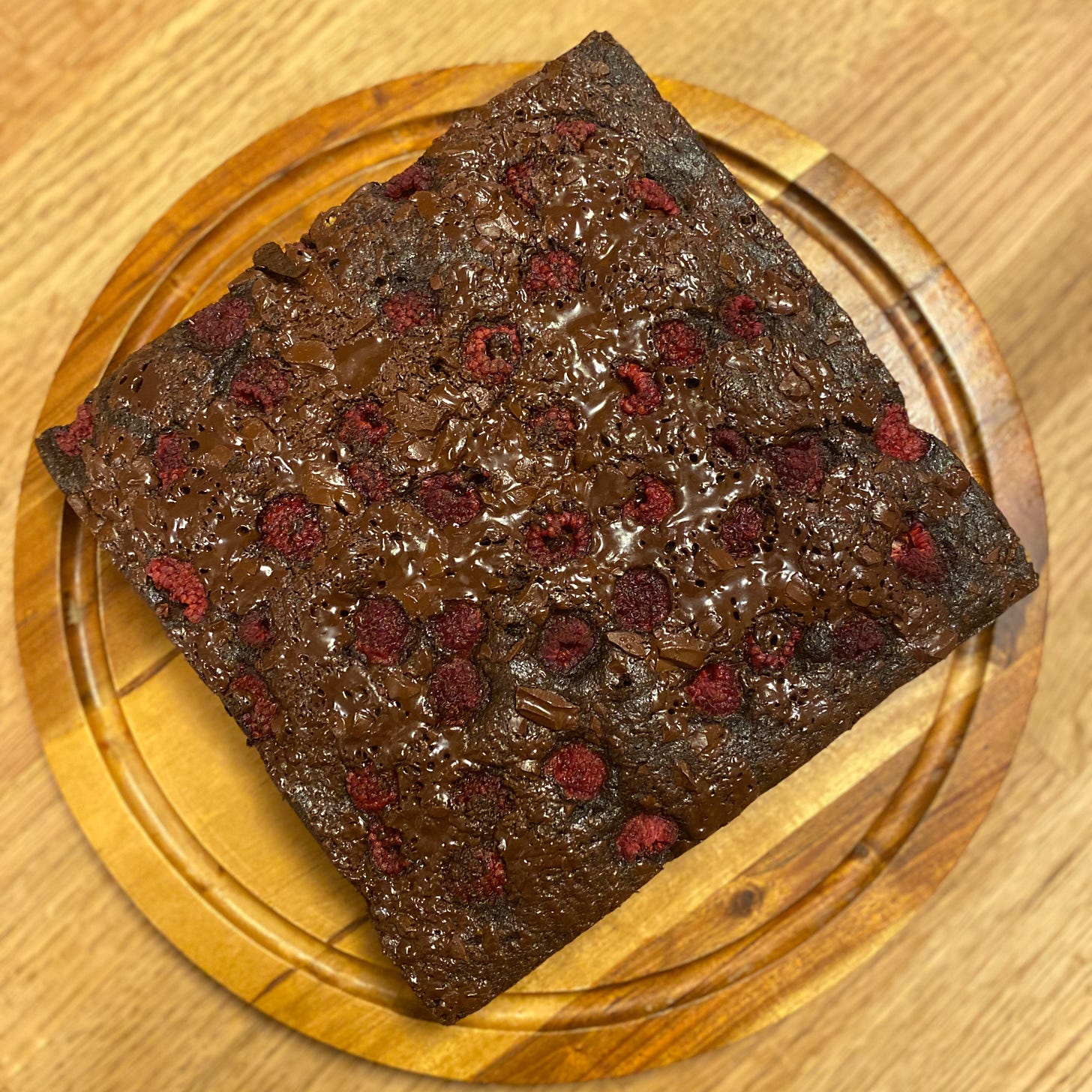 A square chocolate cake, topped with melted chopped chocolate and raspberries, sits on a wooden cake platter.