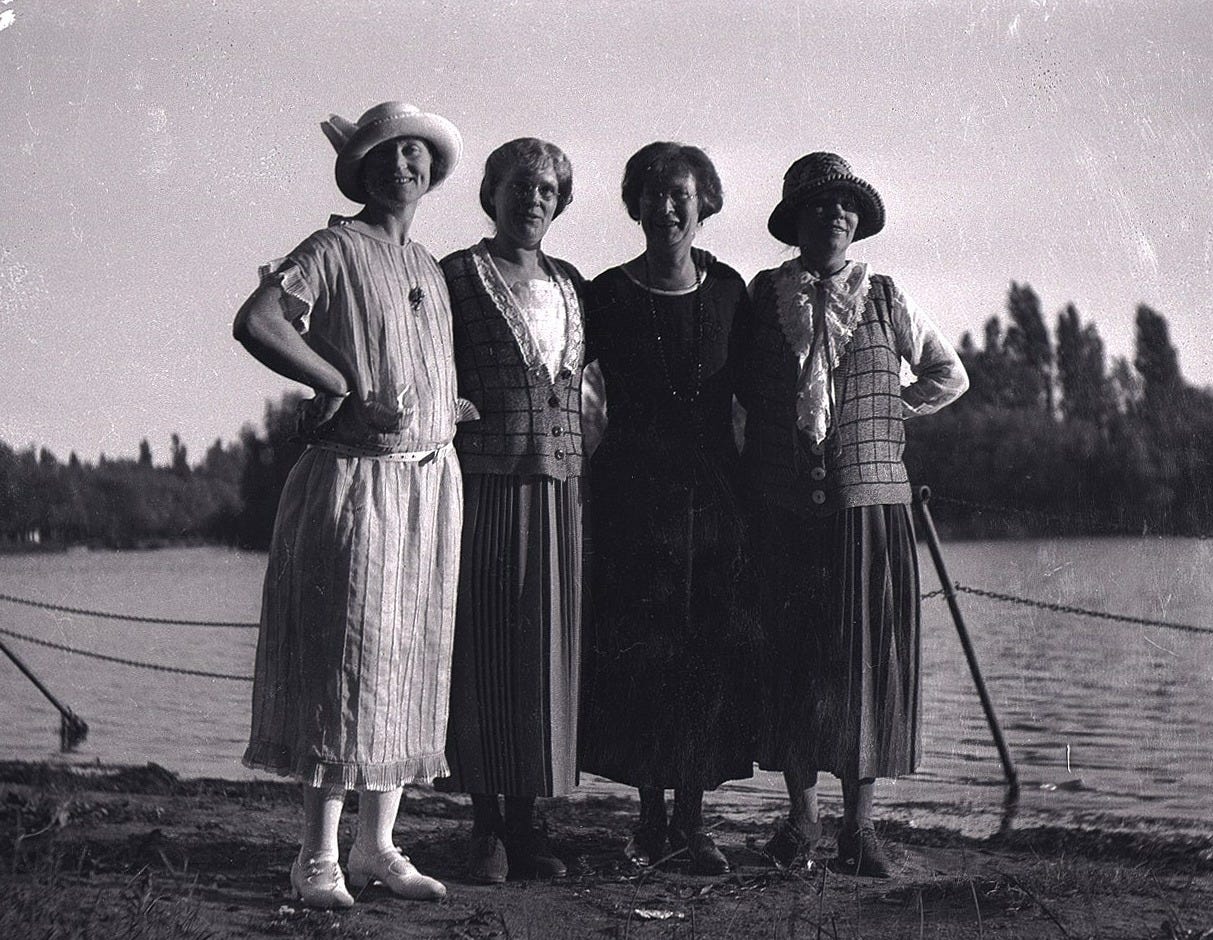 Black-and-white photo of four middle-aged women in 1920s clothing posing in front of a body of water.