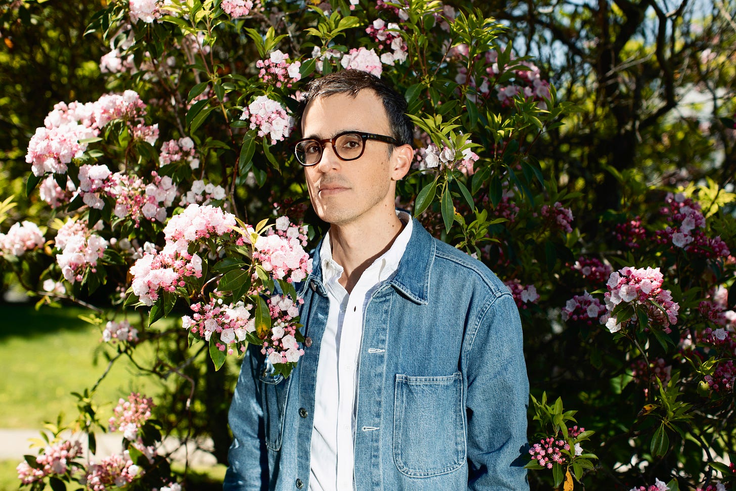 Jonathan Menjivar, wearing a jean jacket, white button down and black frame classes, stands in front of a color bush