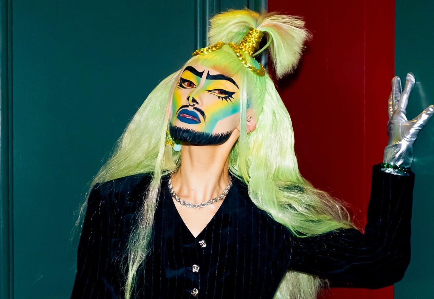 An artist leans against a wall, wearing meticulously painted makeup in yellows, greens, and blues, with a severe painted beard and vibrant green-yellow hair with a lock wrapped in gold thread that shoots straight up at the crown.