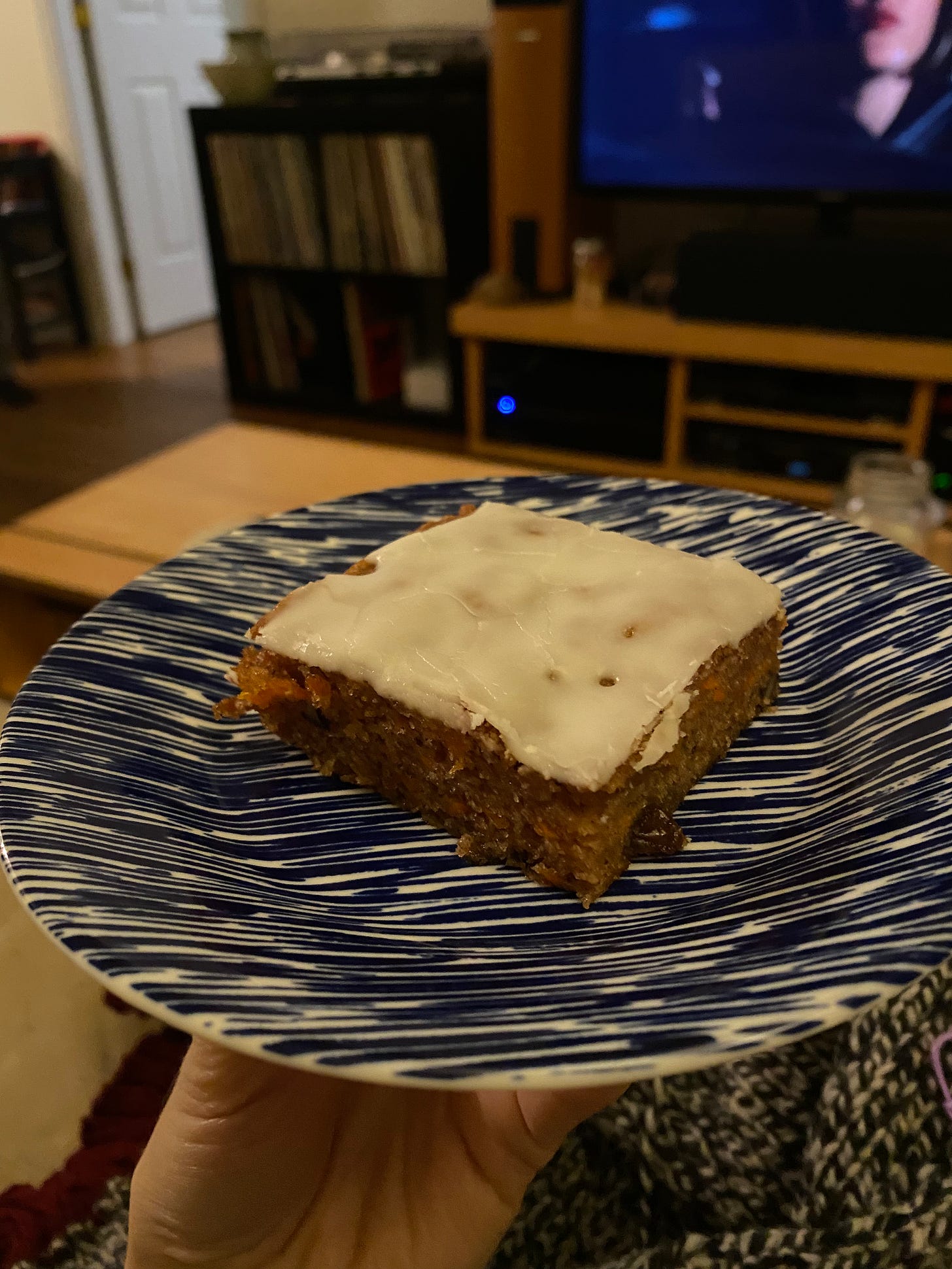 My hand, holding a blue and white plate with a square piece of carrot cake with a thin white icing on top. My knitting is on my lap and you can just see Gillian Anderson on the tv in the background.