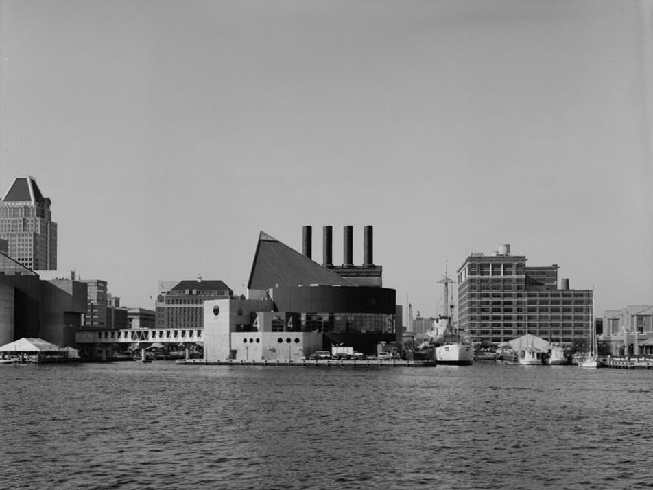 A black-and-white photograph of Baltimore's Inner Harbor redevelopment, prominently featuring the National Aquarium in the center of the image and the smokestacks of the Pratt Street Power Plant behind it.