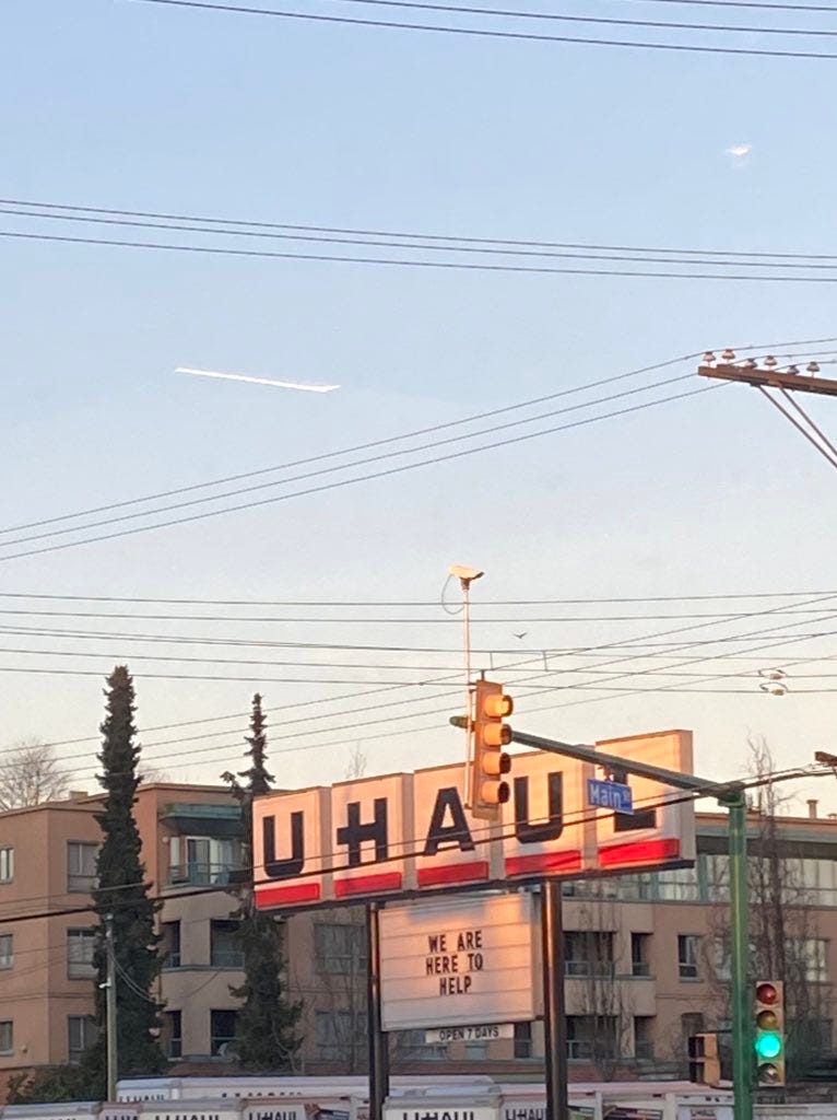 A big U-Haul logo, big block letters in individual signage above a marquee that reads “WE ARE HERE TO HELP” in black text on white. The sky is blue in the background, it’s almost sunset. There are low apartment buildings and the tops of U-Haul vans visible beneath the sign.