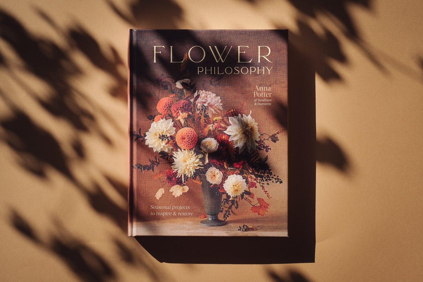 A book titled 'Flower Philosophy' on a paper surface with leaf shadows cast over it. The book cover has a vase with an arrangement of autumn flowers.
