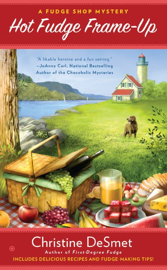Book Cover. Hot Fudge Frame-Up - A Fudge Shop Mystery by Christine DeSmet. Picnic spread with pink fudge and lighthouse and dog in the background.