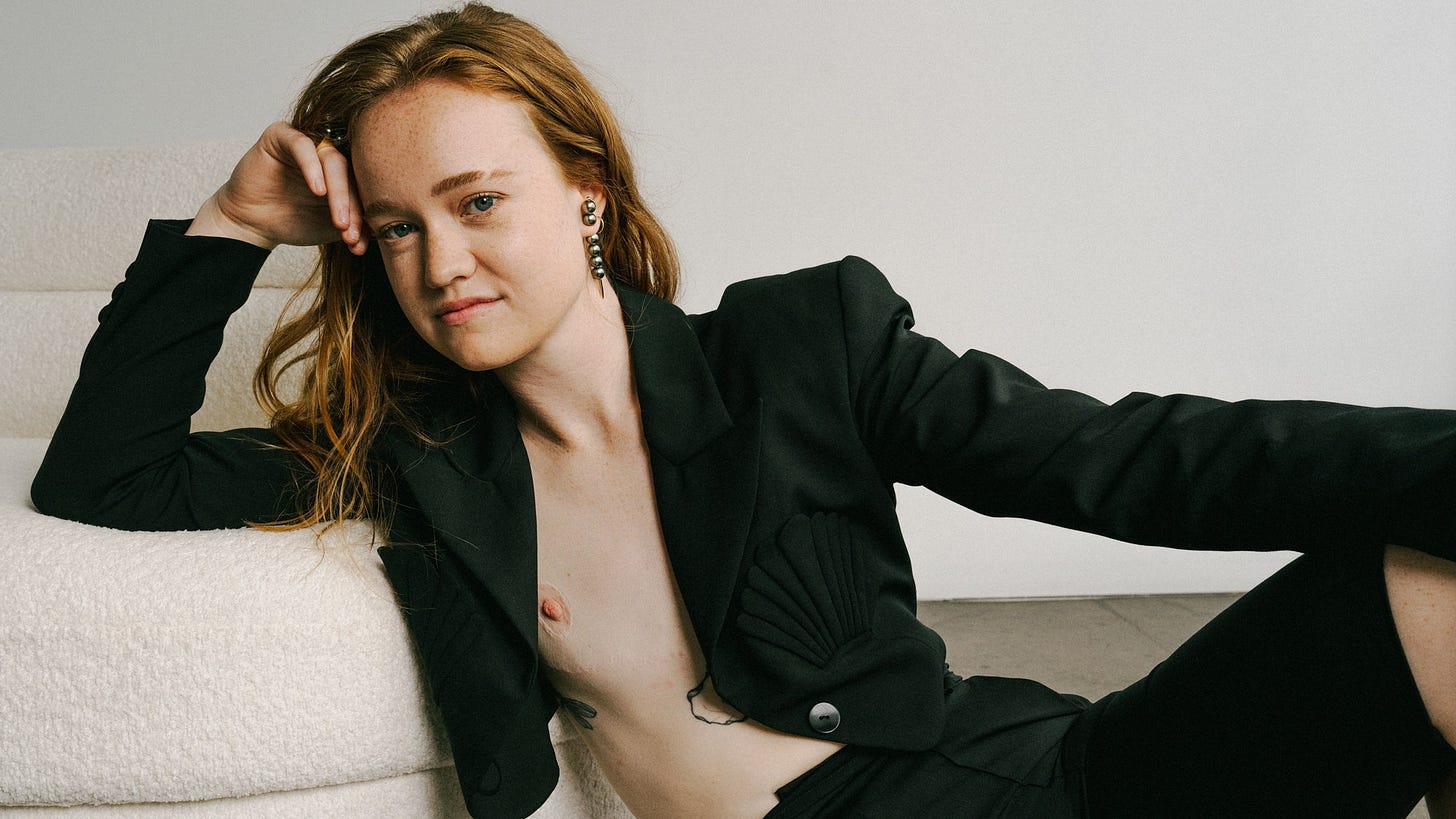 Actor Liv Hewson in a black suit shirtless in front of a white couch