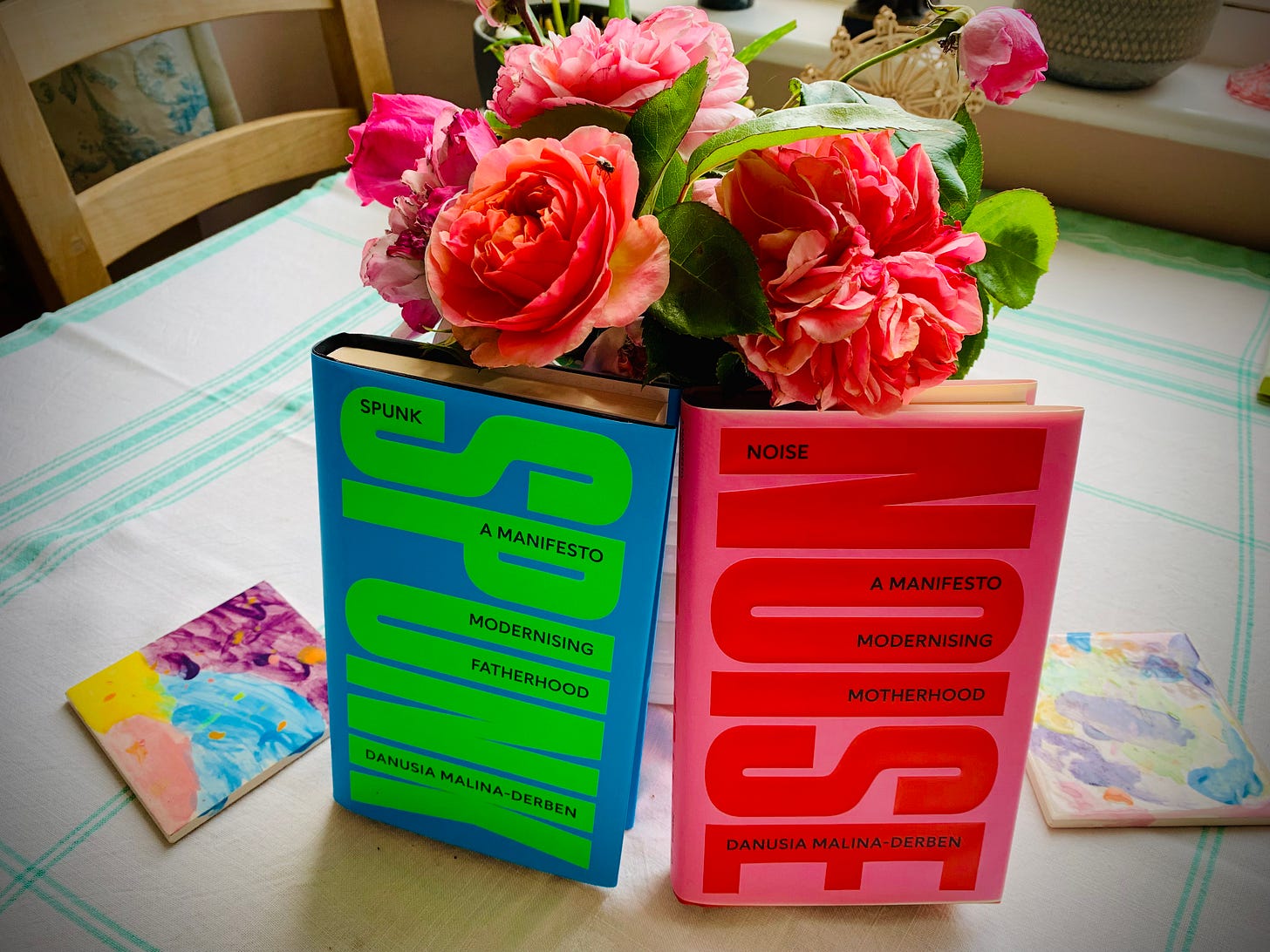 Two brightly coloured hardback books—SPUNK in electric blue and lime green, and NOISE in hot pink and red—stand upright on a kitchen table, nestled against a vase of lush pink roses. Watercolour paintings by children are scattered underneath, evoking warmth, family, and radical rethinking of parenthood.