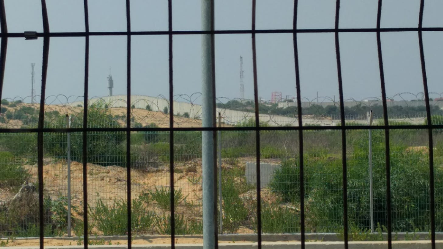 A guard tower tops a long concrete wall, seen through chain link fencing and razor wire