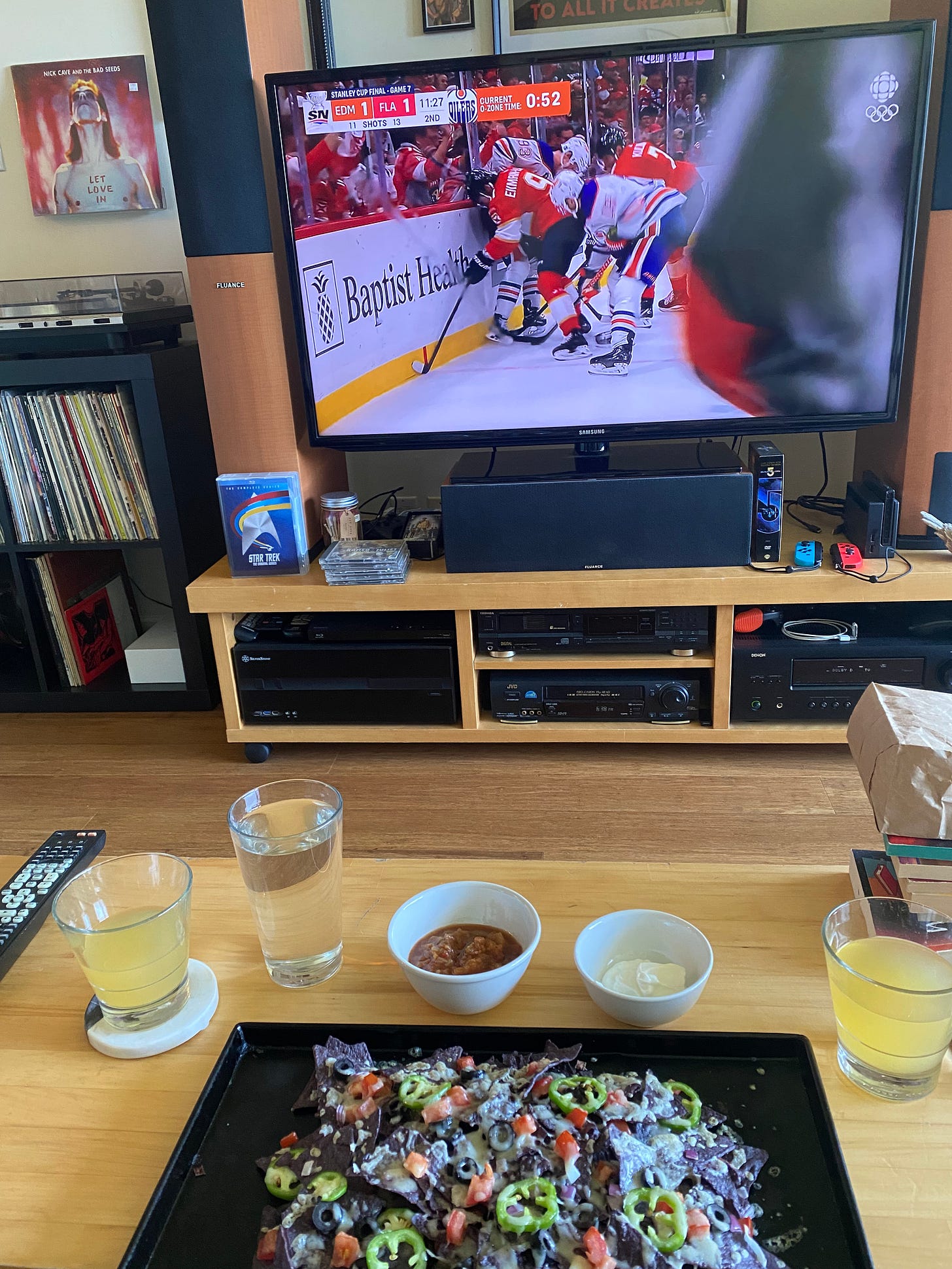 In the background, the hockey game is on the tv. Florida and Edmonton each have one goal. In the foreground, the coffee table has a sheet pan of nachos made with blue corn chips, jalapeño, tomato, and olives. There are bowls of sour cream and salsa behind it, and glasses of beer on coasters to either side.