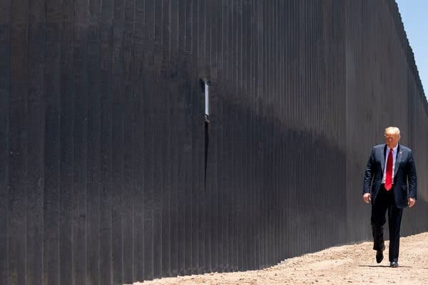 Former President Donald J. Trump walks in a suit and red tie alongside the tall slats of a border fence in Arizona.