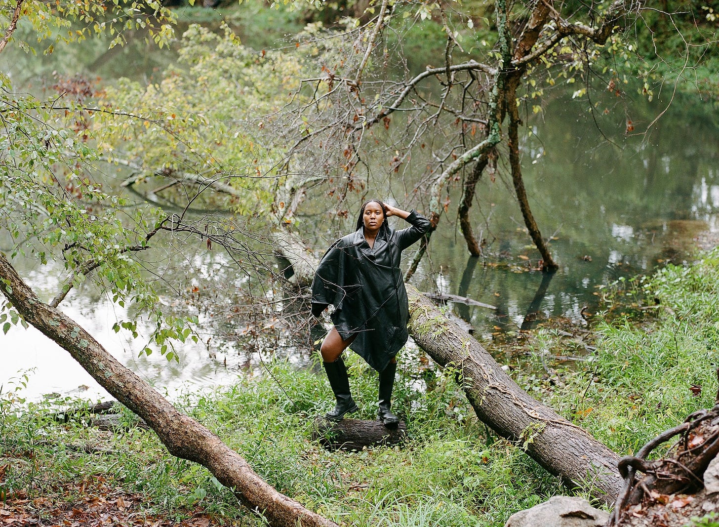 black woman wearing leather jacket/dress standing on a log at the edge of a pond