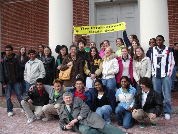 A large group of high school students stand outside a brick building with white columns. An older man in a brown coat lays across the ground in front of them. Two students hold a yellow banner reading The International Brain Bee overhead.