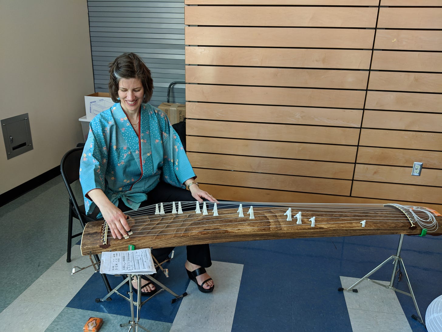 A woman wearing a kimono-style top sits at a koto, or Japanese zither