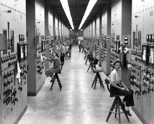 A photo of the calutron girls sitting on benches in front of their machines.