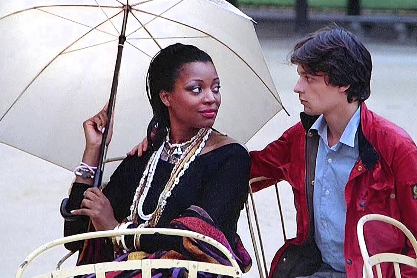 A woman holds an umbrella while sitting next to a man who has his arm around her shoulder.