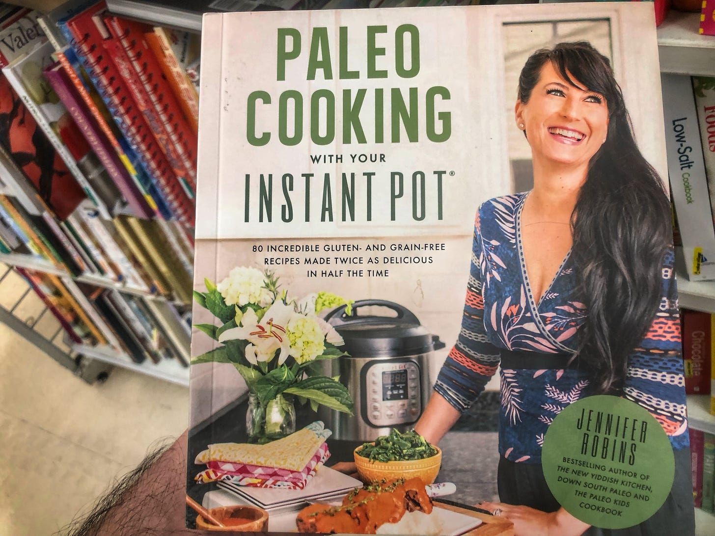 A book called "Paleo Cooking With Your Instant Pot: 80 Incredible Gluten- and Grain-Free Recipes Made Twice as Delicious in Half the Time" by Jennifer Robins, bestselling author of "The New Yiddish Kitchen," "Down South Paleo," and "The Paleo Kids Cookbook" being held up in front of a shelf of cookbooks in a thrift store.