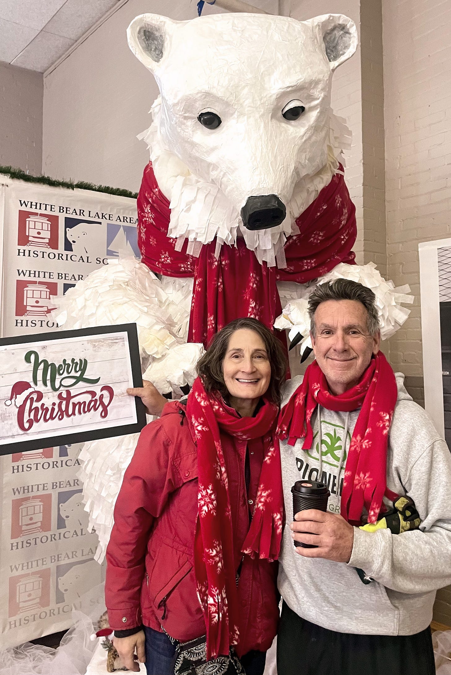 A woman in red winter coat and man with red scarf stand in front of white bear statue with red scarf