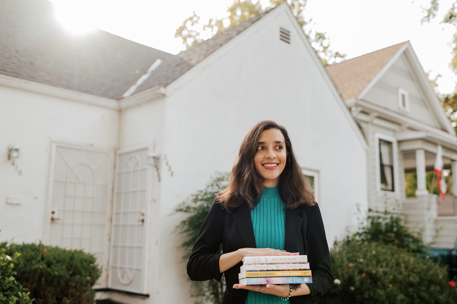 A brown woman with short hair wears a green dress and black blazer. She looks to her left and smiles, holding four books in both hands. She stands in front of a white home. 