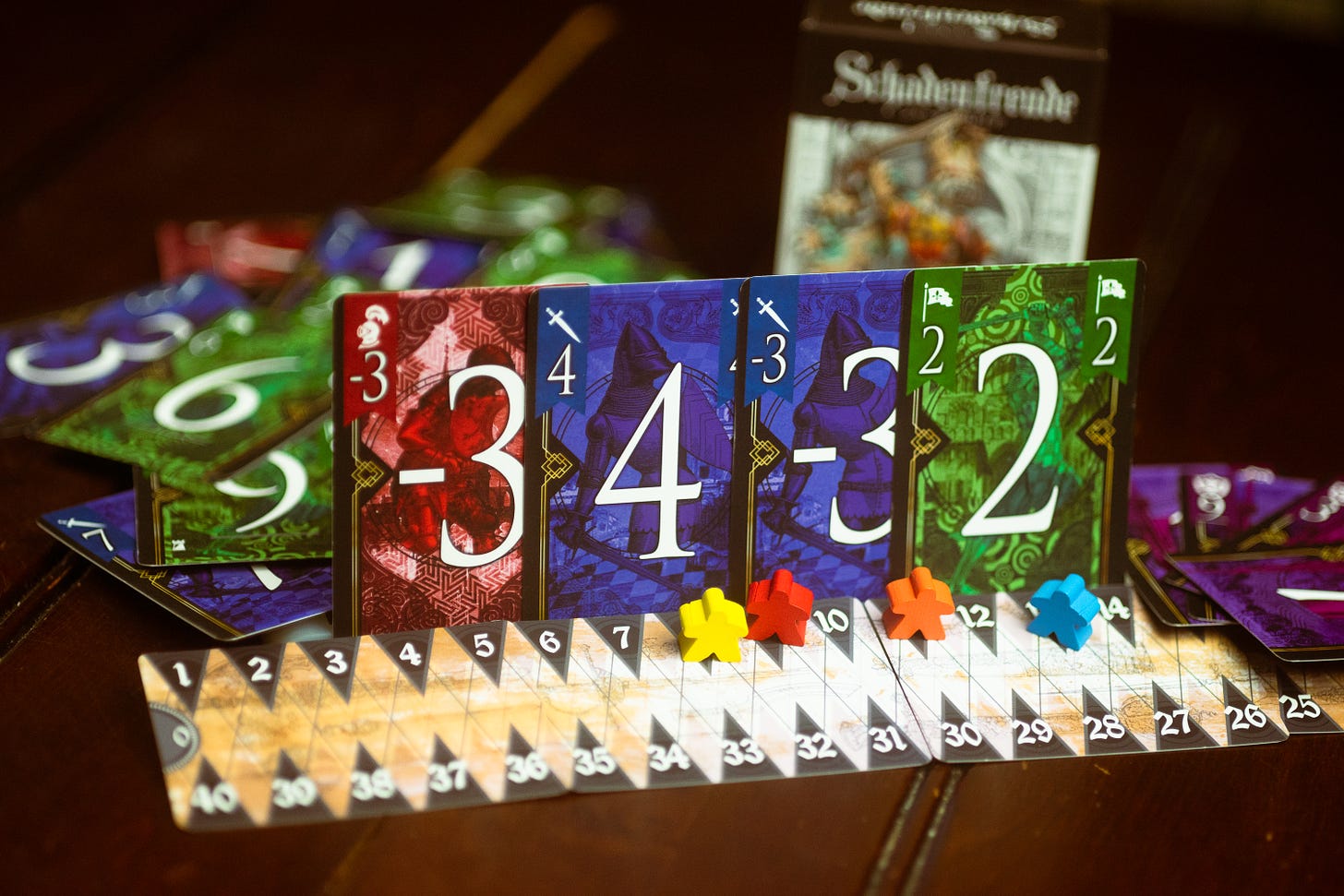 Cards from the game Schadenfreude displayed alongside the scoring track for the game. The box is visible, but out of focus, in the background.