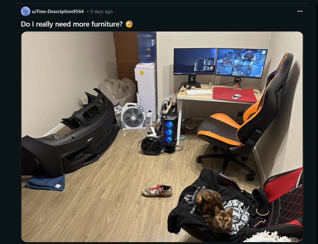 A bare room with a computer desk, a cat in a chair, a fan on the floor, a mini-fridge, and a car bumper.