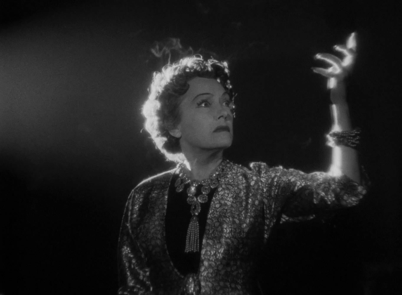 Norma Desmond in Sunset Boulevard standing in her screening room, lit by the projector, holding her hand up like talons.
