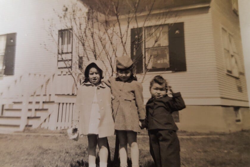 Three young light-skinned children stand holding hands in a black-and-white photo taken circa 1945. Two girls about six years old wear thigh-length coats; a boy about four years old wears a dark suit.