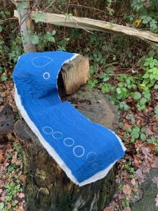 A strip of dark blue fabric, quilted with a design of circles, curves and lines. The piece is backed with grey linen, and rests on a tree stump