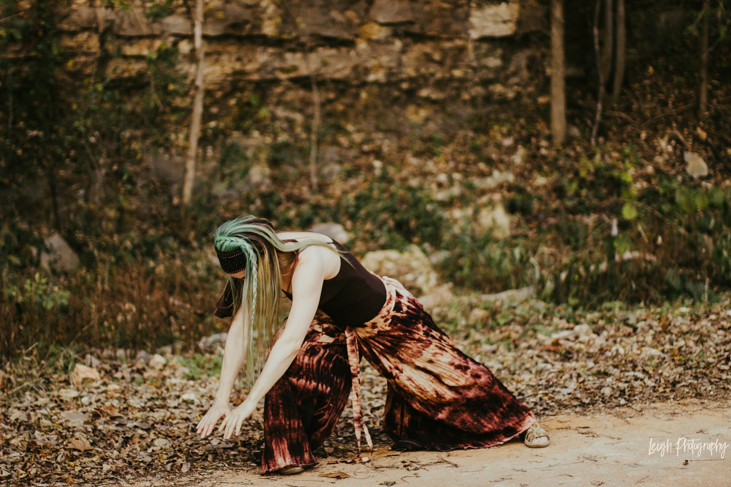 The green-and-brown-haired author in a deep lunge, hands stretched toward the ground. Dancing on the dirt before an immense rock face with shrubs and fallen leaves all around.