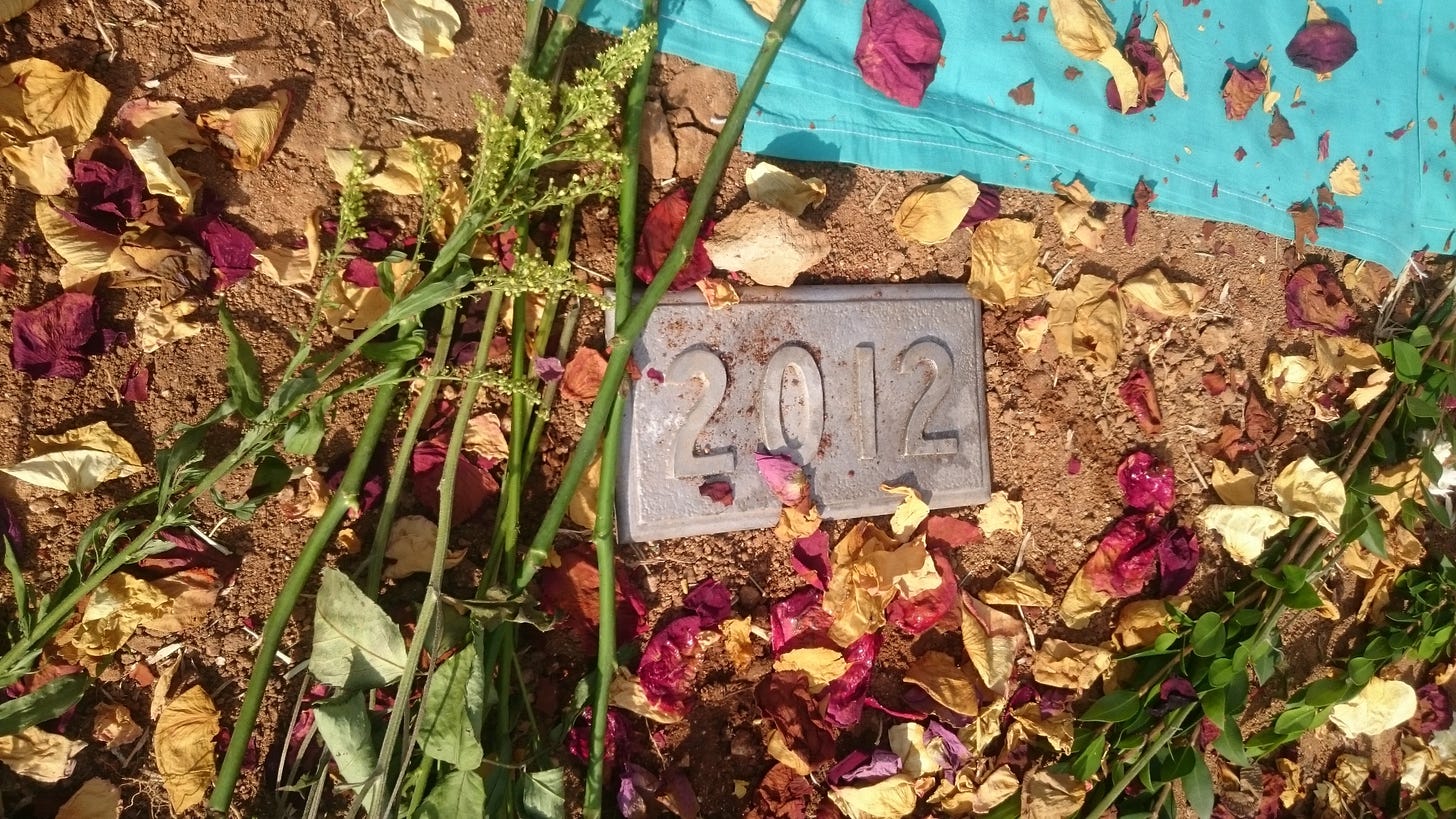 A metal grave marker says only 2012. The marker is surrounded by patches of bare soil and dead flowers