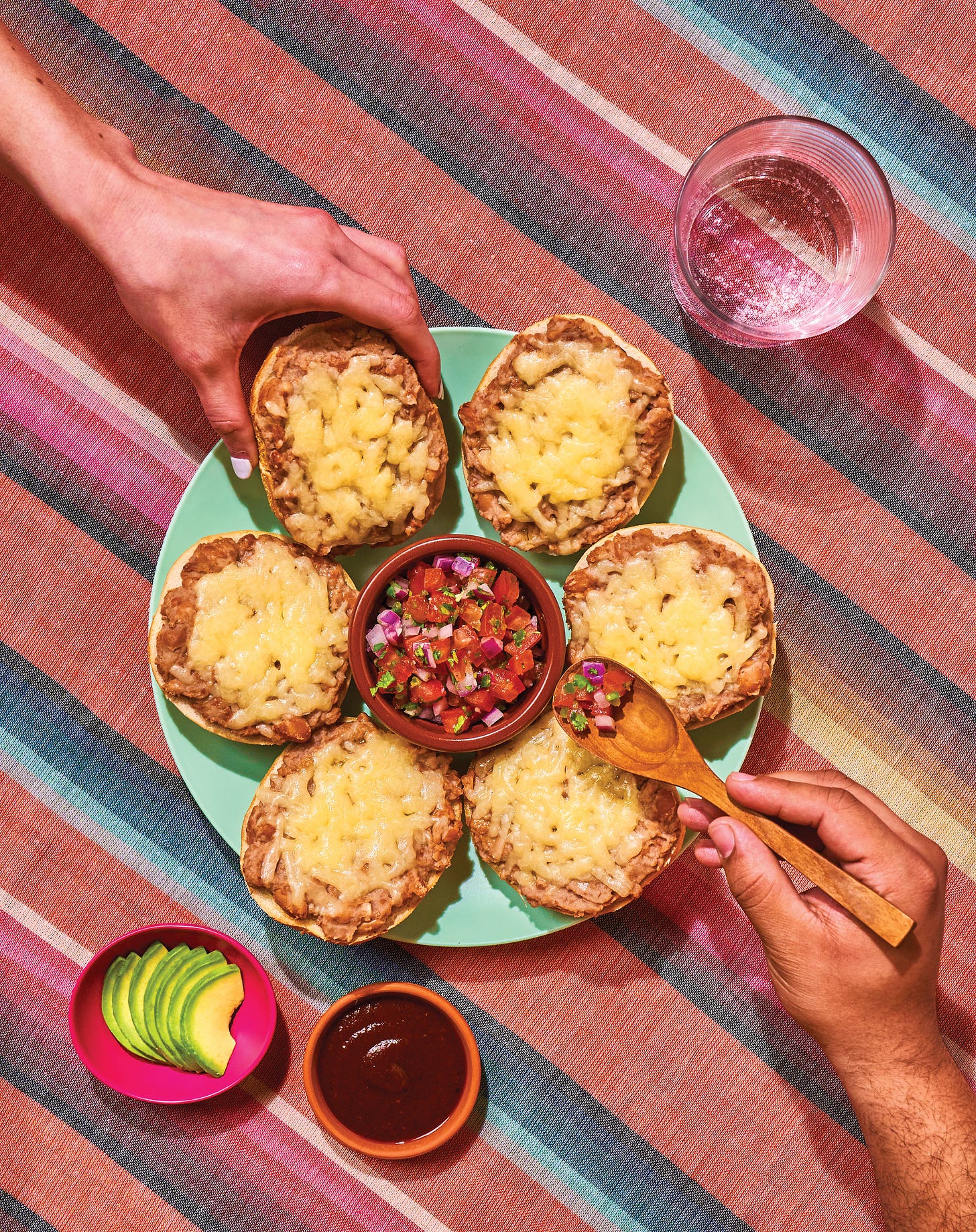 Plate of molletes with pico de gallo, with two hands grabbing, on a striped tablecloth.