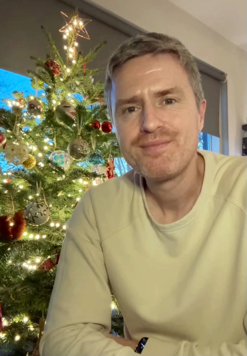 Me sitting in front of my Christmas tree in a cream jumper – my tree is real and has a star at the top and has various glass baubles on it