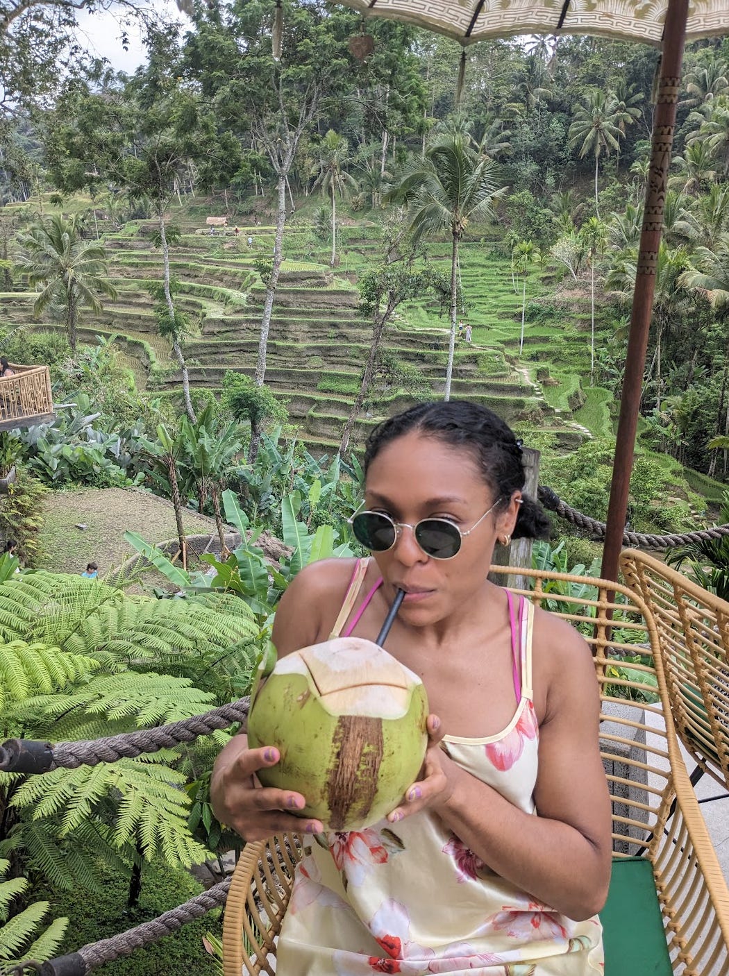 Nathalie sips on a huge coconut in front of hilly rice fields that are visible in the background. She is wearing a yellow dress that has pink flowers on it