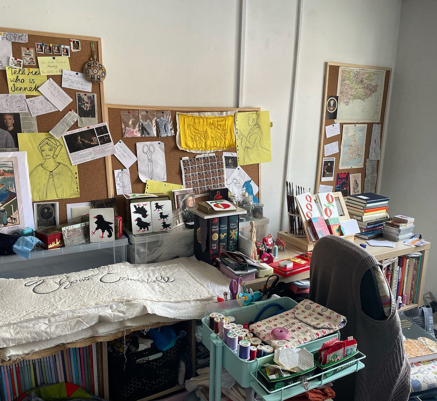 Creative studio view - noticeboards covered in cuttings, notes and maps, a trolley full of stitching supplies, books and a sewing chair. The large Book of Queens artwork is in the foreground.